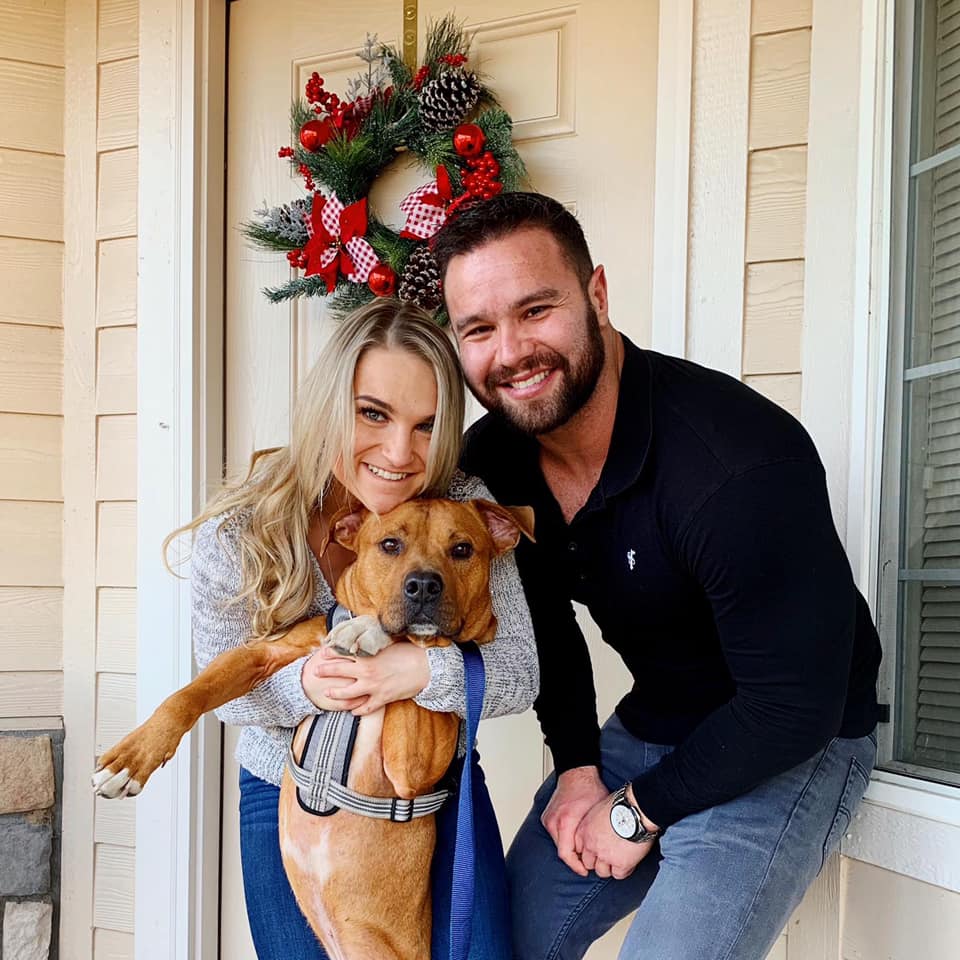 A smiling couple holding a young dog, standing at a home entrance decorated with a festive wreath.