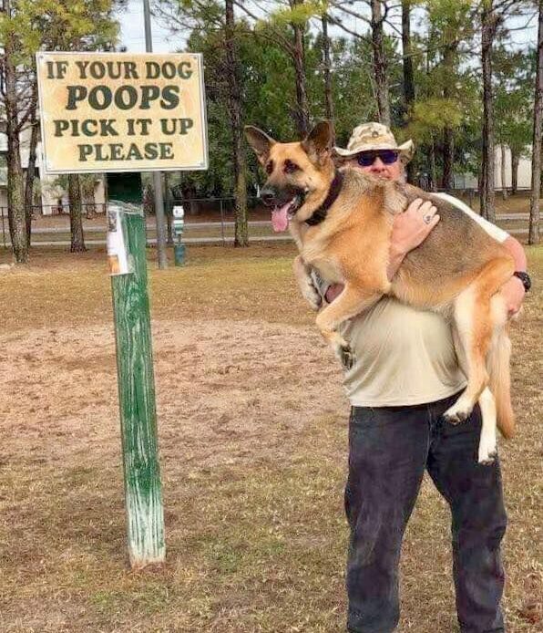 A person humorously carries a German Shepherd dog near a sign instructing dog owners to clean up after their pets.