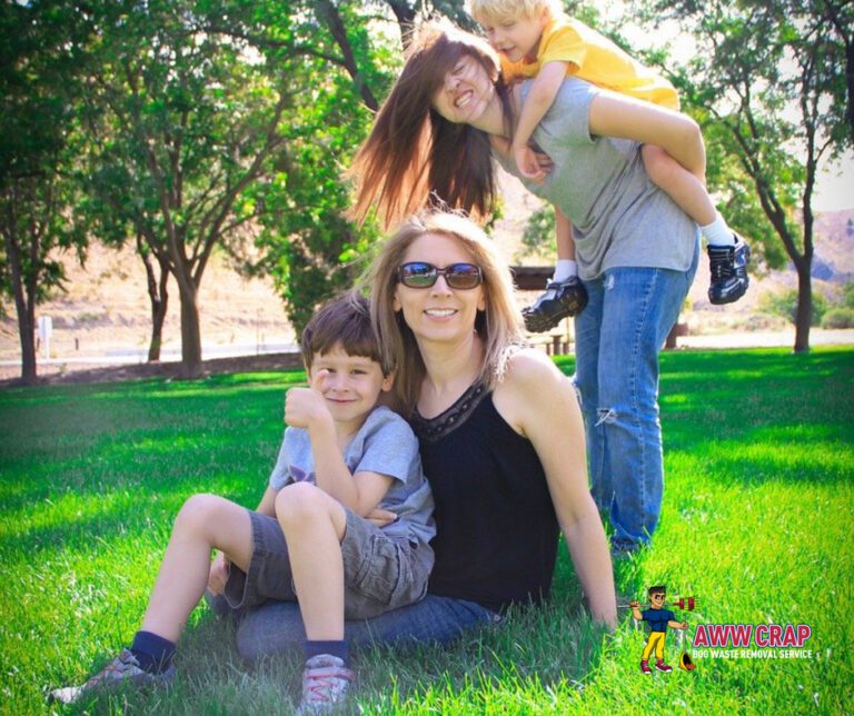 A family enjoying a sunny day outdoors, with two children on their mother's back and another sitting beside her on the grass.
