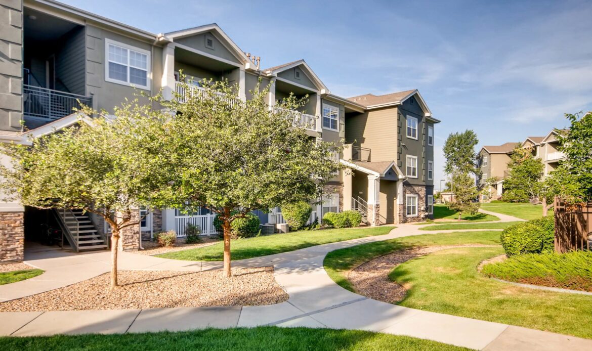 A well-manicured residential area with modern apartment buildings, green lawns, and a clear pathway under a blue sky.