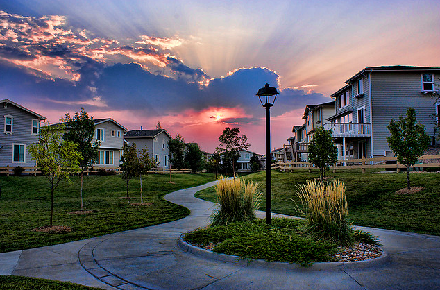 A winding path through a residential area with houses at dusk, under a dramatic sky pierced by sunbeams.