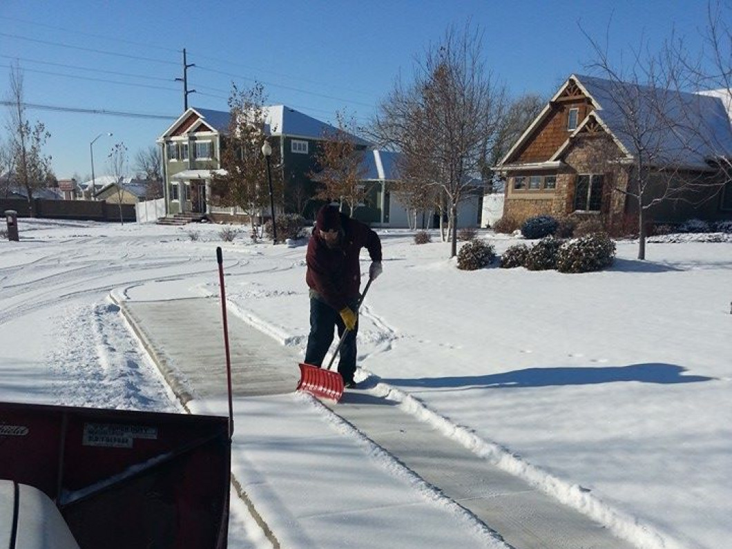 A person is shoveling snow on a sunny day in a residential area with houses in the background.