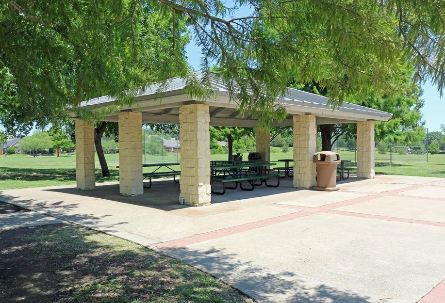 A stone picnic pavilion with tables is nestled under green trees in a sunlit park.