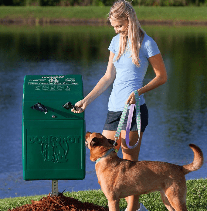 A woman is disposing of dog waste in a designated green bin while her dog stands on a leash beside her.