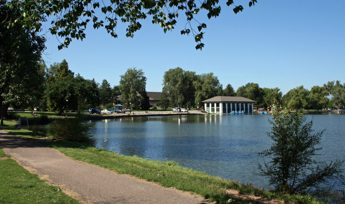 A serene park with a pathway beside a lake, trees, and a building in the distance under a clear blue sky.