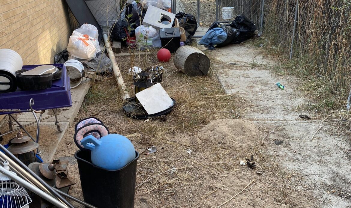A cluttered outdoor area with a chain-link fence, various discarded items, and little vegetation.