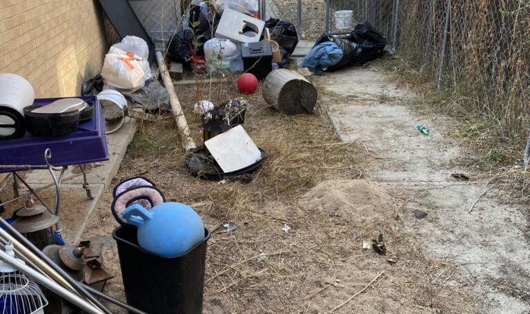 A cluttered outdoor area with a chain-link fence, various discarded items, and little vegetation.