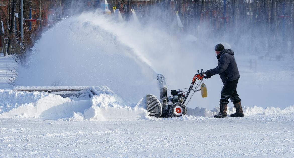 A person operates a snowblower, clearing snow on a sunny day with trees in the background.